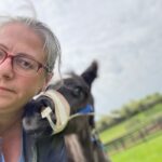 A woman with glasses and a horse in the background.