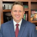 A man in a suit and tie standing next to a book shelf.