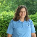 A woman in blue shirt standing next to bushes.